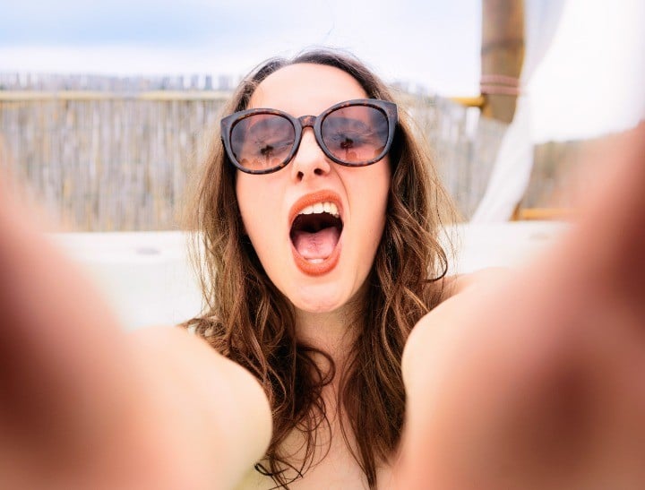 MRMR  S+ A young woman making a funny face as she holds a smartphone to take a selfie on vacation.